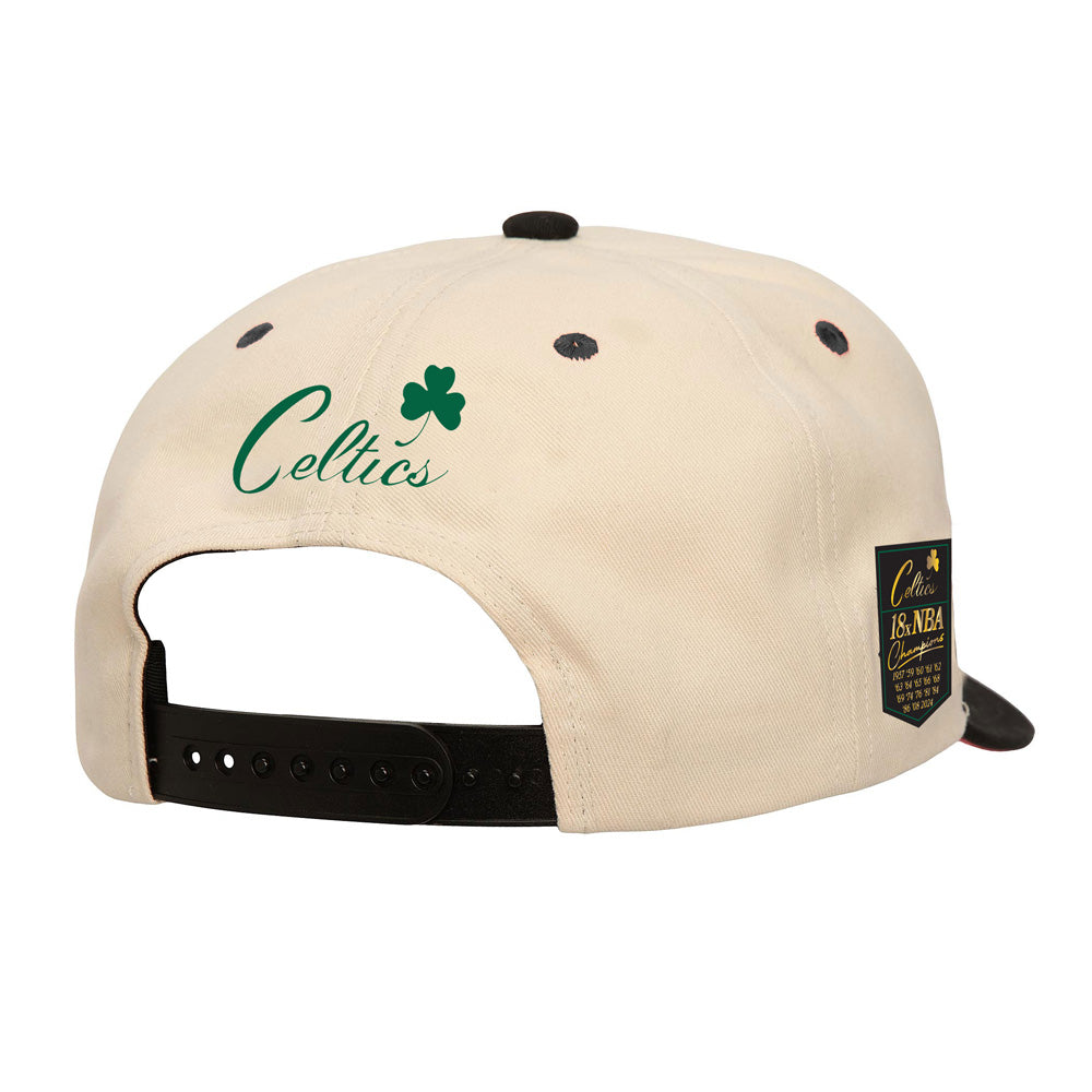 The Mitchell and Ness X Basketball Hall of Fame 18x NBA Champions Boston Celtics cream cap is shown from the back, featuring "Celtics" with a green shamrock above the black adjustable strap, plus a black patch with gold text on the side.