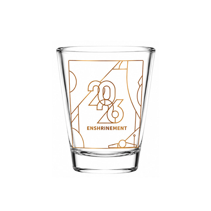 The Basketball Hall of Fame® 2026 Enshrinement Shot Glass features an orange geometric design and "2026 ENSHRINEMENT" on the front. Ideal for basketball collectibles fans. Clear glass on a white background.