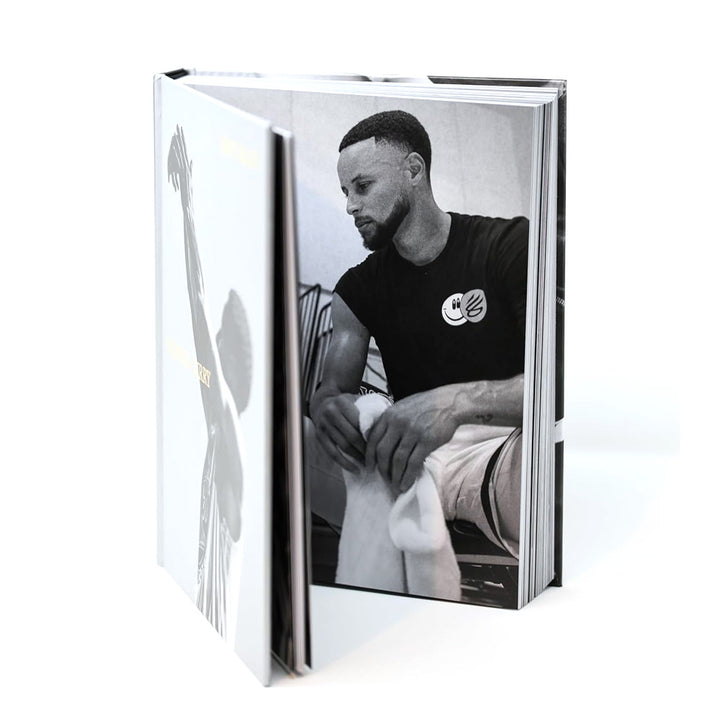 An open hardcover "Shot Ready" book reveals a black-and-white photo of a bearded man sitting with a towel. The cover features a silhouette with one arm raised, capturing the Shot Ready philosophy of success.