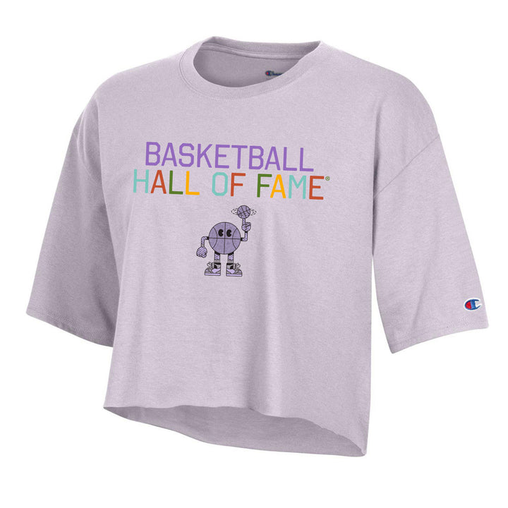The Basketball Hall of Fame® Buckets and Dimes Women's Crop T-Shirt is a light purple crop tee with colorful "BASKETBALL HALL OF FAME" text, a cartoon basketball holding a trophy, and the Champion logo on the left sleeve.