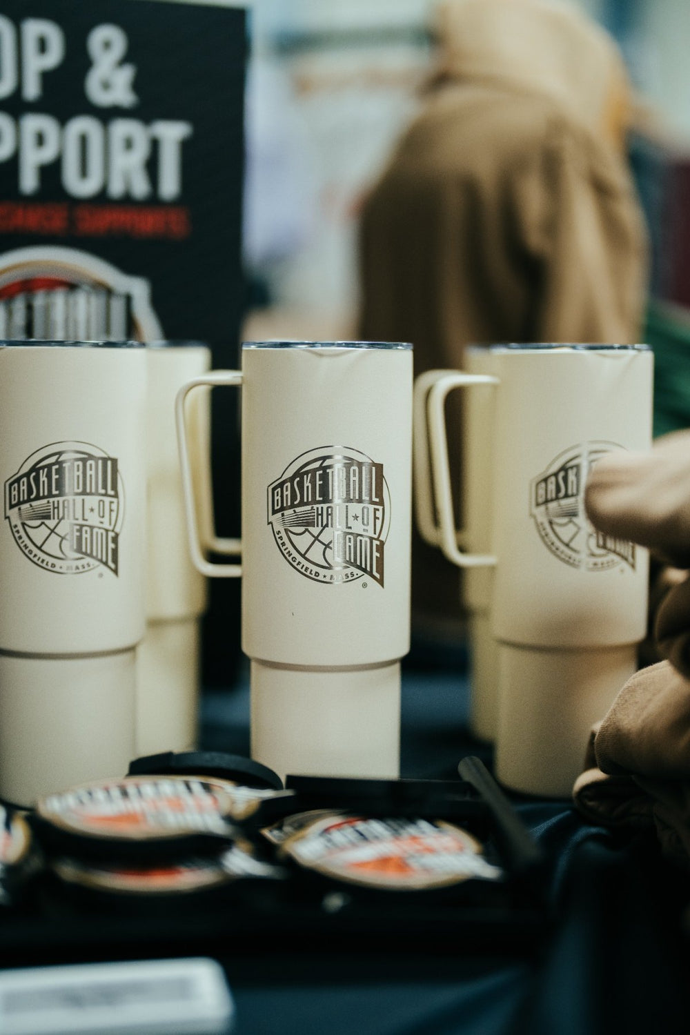 Three cream-colored mugs featuring Basketball Hall of Fame logos are displayed on a table, surrounded by similar logo items such as the Basketball Hall of Fame® 32oz Straw Tumbler and a sports water bottle. A blurred sign and person appear in the background.