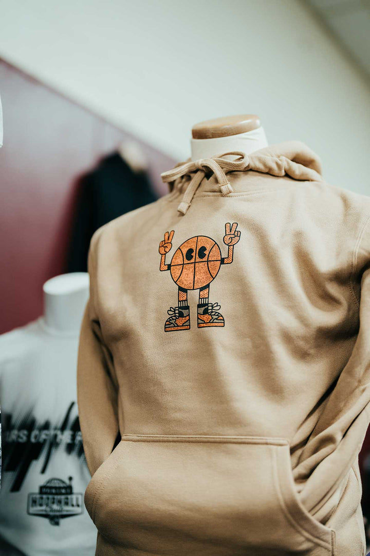 The Basketball Hall of Fame® Basketball Man Hooded Sweatshirt is a tan, unisex hoodie displayed on a mannequin, featuring a playful cartoon basketball character flashing a peace sign. Its sporty design is perfect for basketball enthusiasts and Hall of Fame fans alike, with another graphic white shirt visible in the blurred background.
