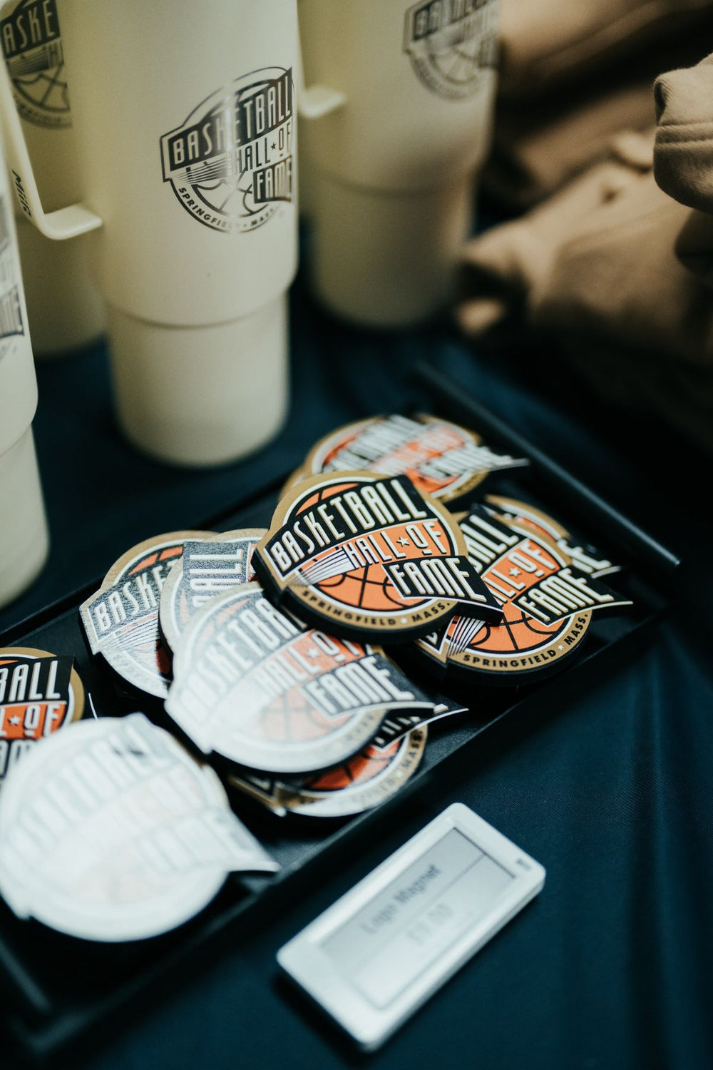 A tray of round “Basketball Hall of Fame®” 3D magnets, perfect as a basketball souvenir, sits next to white tumblers and a small name tag on a dark blue tablecloth.