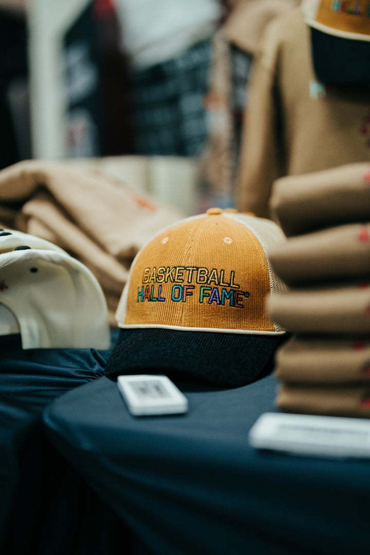 A Basketball Hall of Fame® Cord Trucker Hat, featuring a yellow and black cord design with embroidered detailing, sits on a table near stacks of folded brown shirts and other caps in the background—making it the perfect accessory for any basketball enthusiast.
