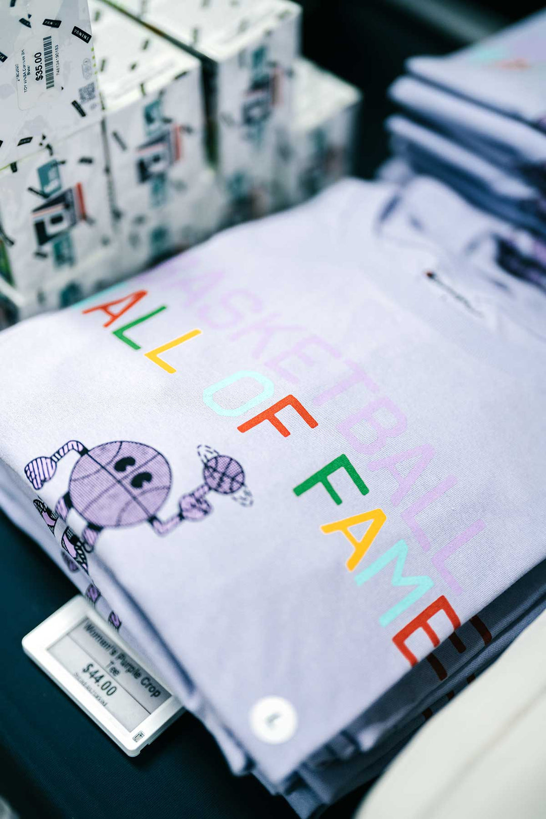 A Basketball Hall of Fame® Buckets and Dimes Women's Crop T-Shirt in lavender, featuring a cartoon basketball holding a diamond and the text "BASKETBALL ALL OF FAME" in colorful letters, is displayed on a table next to stacked boxes. Perfect for basketball enthusiasts or anyone inspired by the legacy of the Hall of Fame. Price: $44.00.