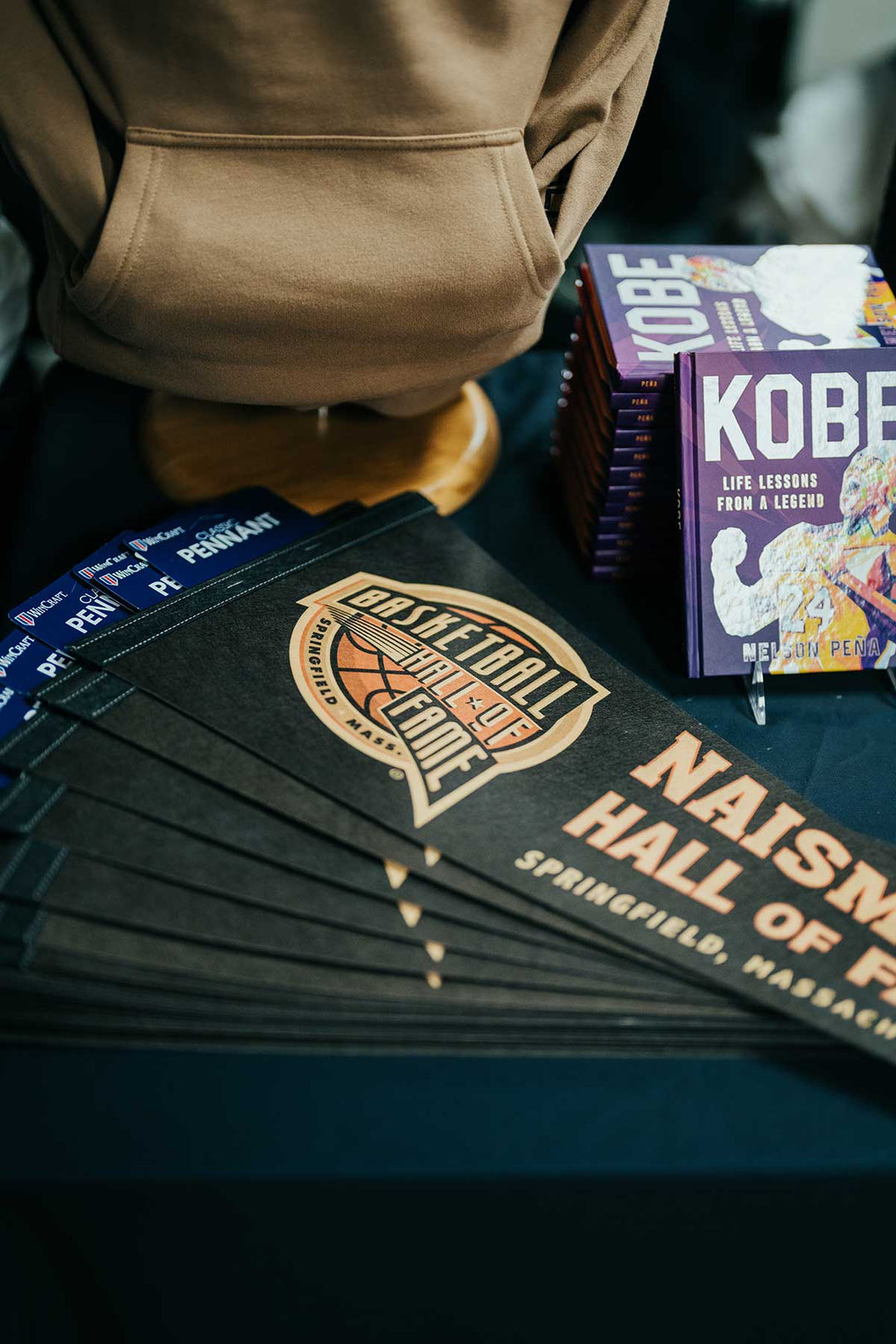 A stack of Classic Basketball Hall of Fame Pennants and a pile of Kobe books are displayed on a dark table next to a brown hoodie, creating a striking arrangement of basketball memorabilia.