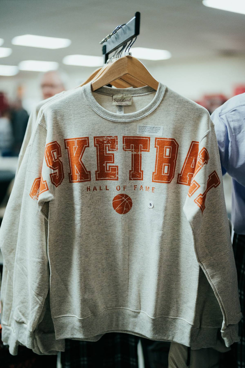 The Basketball Hall of Fame® Spread Arms Fleece Sweatshirt is a gray sweatshirt displayed on a hanger, featuring "BASKETBALL HALL OF FAME" and a basketball graphic printed in bold orange and white letters—perfect for fans who appreciate basketball history.