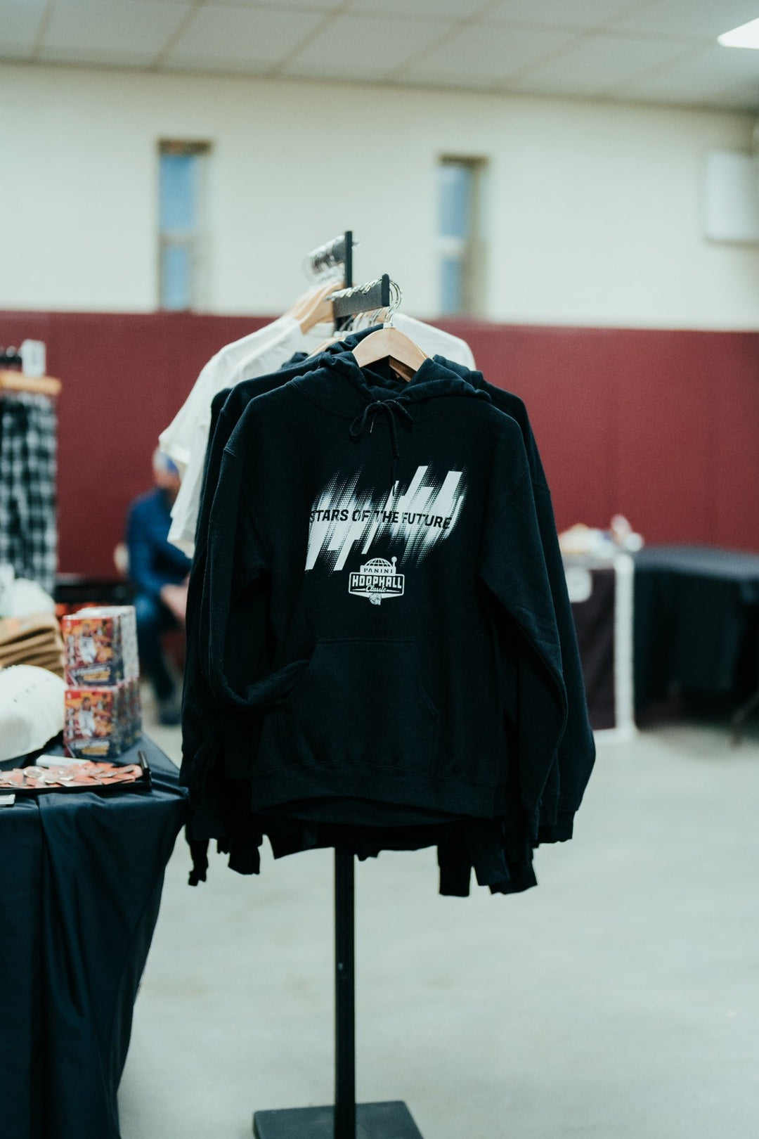 A 2026 Hoophall Classic Hooded Sweatshirt, black with "STARS OF THE FUTURE" printed on the front, hangs on a rack in an indoor market setting, capturing the spirit of the Panini Hoophall Classic. Other clothing items are partially visible nearby.