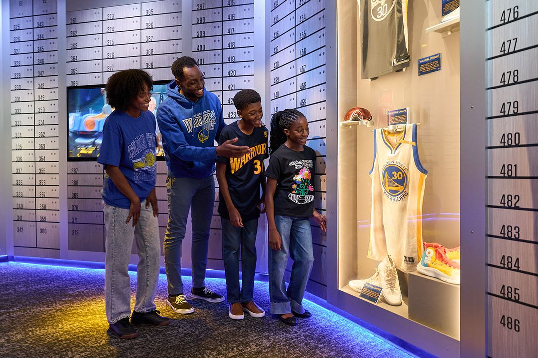 A man wearing an NBA Mitchell & Ness Golden State Warriors Hooded Sweatshirt and three children look at a jersey and shoes displayed in a glass case at a museum.